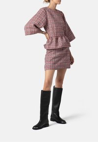 Mujer vistiendo un traje de falda a cuadros en tonos rosa y gris, con una blusa tipo peplum y botas de cuero negras hasta la rodilla, de pie con una mano en la cadera.