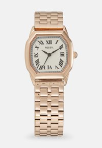 Montre octogonale en or rose Fossil avec un cadran blanc texturé, des chiffres romains noirs et un bracelet en maillons métalliques. Elle présente un fermoir simple.