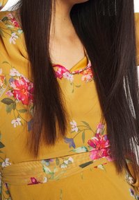 Blus i blomstermönster i gult med rundad halsringning, prydd med rosa och vita blommor samt en strukturerad, lagerdesign vid midjan.