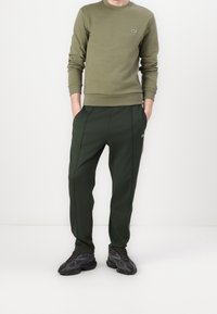 Sudadera verde oliva con escote redondo y puños de canalé, acompañada de pantalones oscuros verde ajustados con bolsillos laterales y un pequeño logotipo.