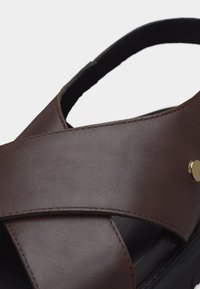 Καφέ δερμάτινο σανδάλι με φαρδιές λουρίδες που διασταυρώνονται και μαύρη μαλακή σόλα, με ένα μικρό στρογγυλό χρυσό κουμπί στο πλάι.