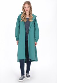 Zöldesszürke hosszú esőkabát kapucnival, rugalmas mandzsettákkal, elülső cipzárral és oldalzsebekkel, csíkos ing és farmer fölött viselve, sportcipővel párosítva.