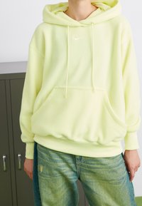 Sudadera de forro polar en amarillo claro con cordones, bolsillo delantero y logo bordado. Combinada con vaqueros azules desgastados.