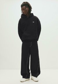 Jeune personne avec des dreadlocks portant un hoodie noir, un pantalon noir ample et des baskets blanches, se tenant contre un fond léger uni.