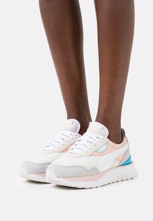 Personne portant des baskets Puma blanches et rose pastel avec des accents bleus, debout sur une surface blanche contre un fond uni.