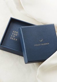 Marineblauwe geschenkdoos met een gestructureerd oppervlak, voorzien van gouden tekst "ZIE HET ONGEZIENE" en "VIOLET HAMDEN," evenals een accent van een halve maan. De doos is gedeeltelijk geopend.