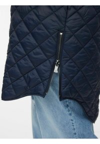 Gilet matelassé marine avec un ourlet asymétrique et des détails de fermeture éclair argentée, présentant une texture douce et un motif en losanges ; superposé sur un denim clair.