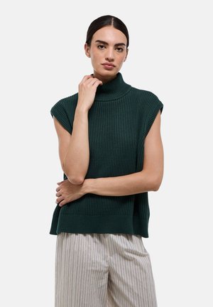 Pull à col roulé sans manches vert foncé avec une texture côtelée. Les fentes latérales améliorent la circulation de l'air. Le modèle porte un pantalon à rayures larges et de couleur claire.