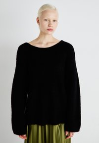 Black knit sweater with a wide neckline and long sleeves, featuring a textured, soft fabric. Worn over a green pleated skirt.