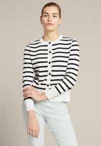 Gestreifter schwarzer und weißer Cardigan mit Rundhalsausschnitt, Knopfverschluss und gerippten Bündchen. Über einer hellen Hose getragen. Glatte Stricktextur.