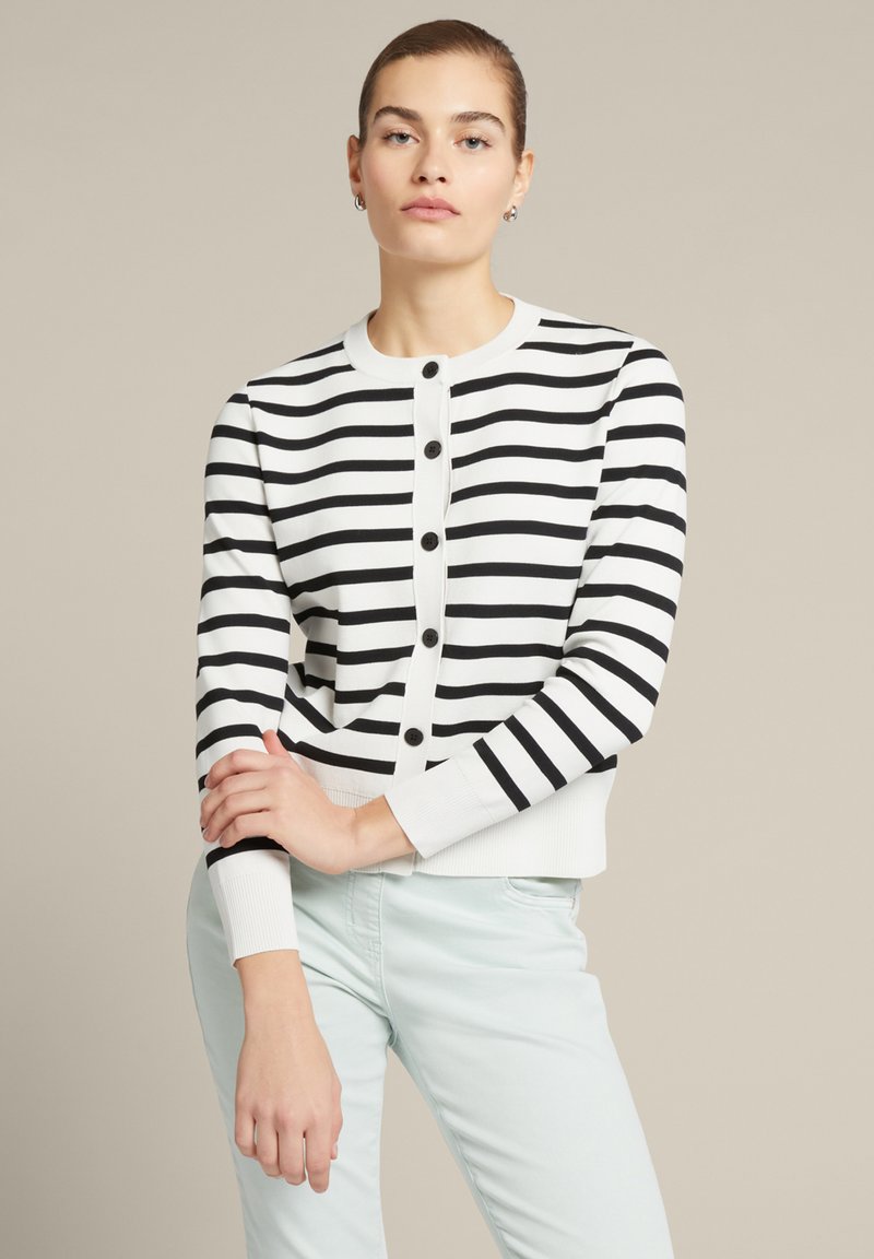 Gestreifter schwarzer und weißer Cardigan mit Rundhalsausschnitt, Knopfverschluss und gerippten Bündchen. Über einer hellen Hose getragen. Glatte Stricktextur.
