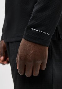 Κοντινό πλάνο ενός σκούρου χρώματος δέρματος χεριού που φοράει μαύρο πουκάμισο με μακριά μανίκια, με την επιγραφή "OMNI-FREEZE" κοντά στον καρπό.