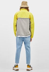 Värilohkottu takki, jossa keltaiset hihat ja harmaa runko; siinä on korkea kaulus ja joustavat hihansuut. Yhdistetty vaaleansinisiin housuihin ja monivärisiin tennareihin.