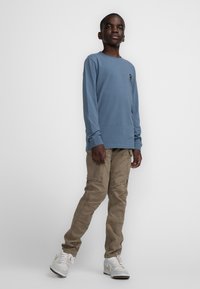 Lange mouwen blauwe t-shirt met een klein logo, gecombineerd met beige cargobroek met meerdere zakken en witte sneakers met gouden accenten.