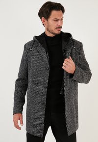 Schwarz-weiße gemusterte Jacke mit Kapuze, Knopfschluss, seitlichen Taschen und kontrastierendes schwarzes Innenfutter. Hergestellt aus strukturiertem Stoff.