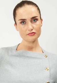 Harmaa neule, jossa on veneen kaula-aukko ja kultaiset napit yhdellä olalla. Kangas näyttää pehmeältä ja sileältä.