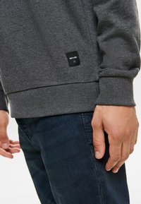 Grijze katoenen sweatshirt met ribgebreide boorden en een onderkant, voorzien van een zwart logo patch. Draagt over donkerblauwe spijkerbroek, hand aan de zijkant geplaatst.