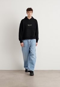 Schwarzer Kapuzenpullover mit grafischen Details, lockere hellblaue Jeans und schwarze Slipschuhe. Glatte Textur, lässiges Design, entspannte Passform.