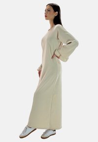 Vestido beige de manga larga con un corte relajado, textura acanalada y puños fruncidos, combinado con zapatillas blancas con suelas marrones.