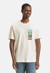 Cremefarbenes Baumwoll-T-Shirt mit kurzen Ärmeln, mit einem rechteckigen grafischen Druck in Grün und Braun auf der linken Vorderseite.