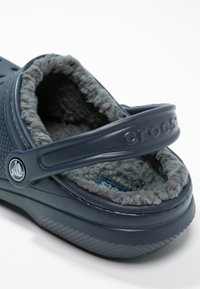 Chausson en caoutchouc bleu foncé avec une doublure grise duveteuse, design moulé, bride arrière et un détail de logo circulaire sur le côté.