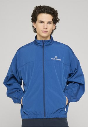 Jonge man met krullend haar, gekleed in een blauwe Sergio Tacchini jas met rits, met handen in de zakken tegen een effen achtergrond.