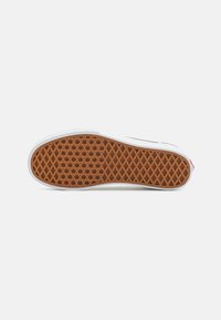 Suela de zapatilla blanca con una parte inferior de goma marrón texturizada que presenta un patrón de rejilla en forma de diamante. Bordes blancos lisos contornan la suela.