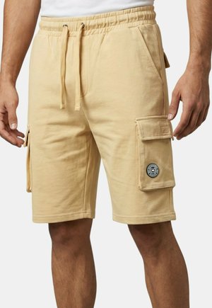 Shorts cargo beiges avec taille à cordon, poches latérales, et un écusson rond noir et blanc sur la poche cargo droite, portés par une personne debout.