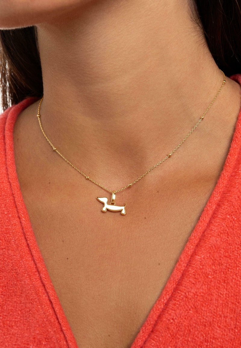 Goldkette mit einem Dackel-Anhänger, mit einem polierten Finish, vor einem korallenfarbenen Oberteil.