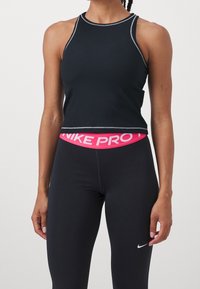 Mujer vistiendo un top corto sin mangas negro y leggins atléticos de cintura alta negros con una cinturilla rosa de Nike Pro y el logo blanco de Nike.