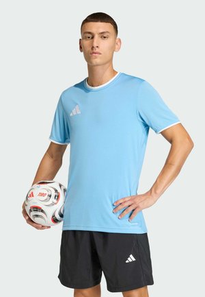 Młody mężczyzna w jasnoniebieskiej sportowej koszulce i czarnych szortach trzyma piłkę nożną jedną ręką, stojąc z drugą ręką na biodrze.