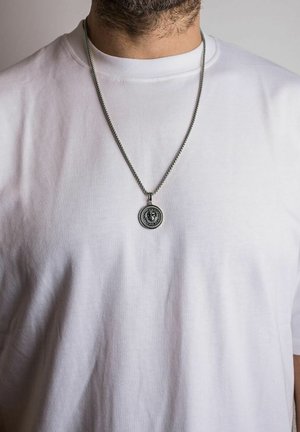 Silver chain necklace with a circular lion medallion pendant. Worn over a plain white t-shirt.