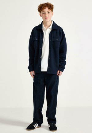 Ragazzo dai capelli rossi in piedi, indossa una giacca blu navy, camicia bianca, pantaloni blu navy e scarpe da ginnastica nere con strisce bianche su uno sfondo semplice.