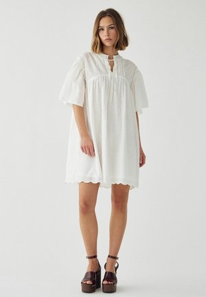 Mladá žena stojaca, oblečená v voľných bielych šatách s nafúknutými rukávmi a okrúhlym lemom, doplnených tmavohnedými platformovými sandálmi.