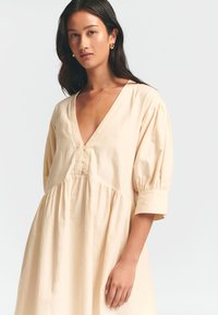 Beige Baumwollkleid mit tiefem V-Ausschnitt, Puffärmel und Knopfdetails an der Brust. Verfügt über einen fließenden Rock mit gerafftem Bund.