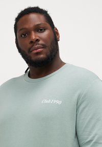 Lysegrøn sweatshirt med rund hals og broderet "Club1990" detalje på brystet, lavet af blødt bomuldsmateriale.