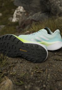 Chaussures de course bleu clair et blanc avec semelles en caoutchouc noires Continental placées sur un terrain rocheux et herbeux.