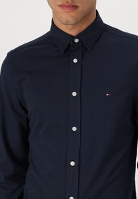 Camisa azul marino de botones con un patrón de espiga texturizado, con botones blancos y un pequeño logotipo en el pecho izquierdo.