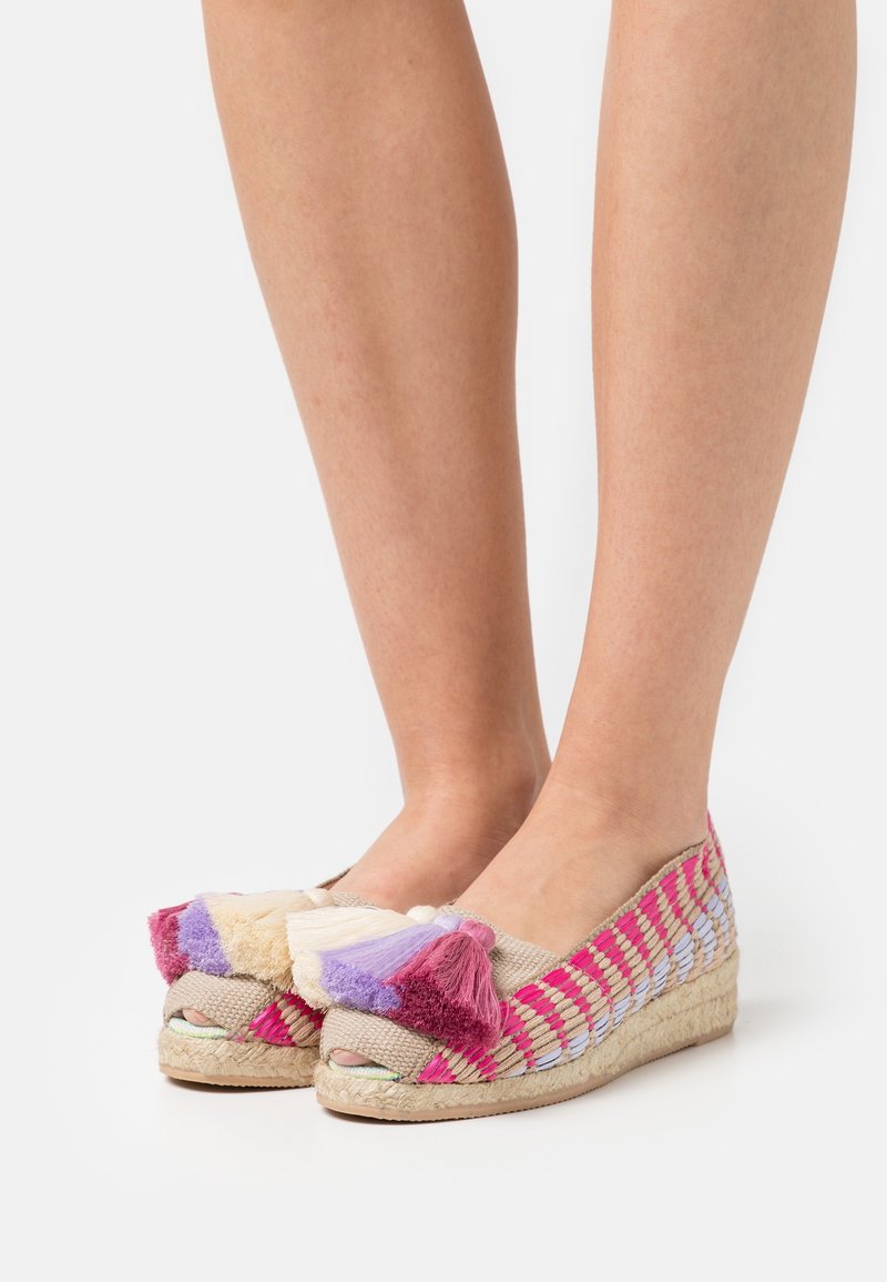 Pieds portant des chaussures compensées tissées rose et beige avec des pompons multicolores sur le dessus, sur un fond blanc.