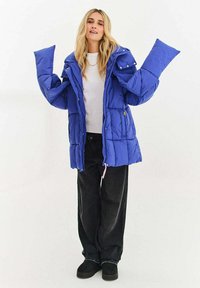 Modrá prošívaná bunda s nadměrnými rukávy, se zipem a zapínáním na druky, bočními kapsami a texturovaným povrchem. Nositelná přes bílý top a černé kalhoty.