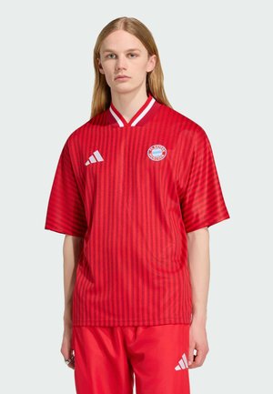 Osoba z długimi włosami ubrana w czerwoną koszulkę FC Bayern Monachium i czerwone szorty Adidas na jednolitym jasnym tle.
