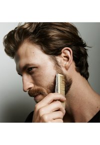 En person använder en metallkam med ett dekorativt handtag för att styla sitt skägg. Kammen har långa, fina tänder och personens hår är brunt.