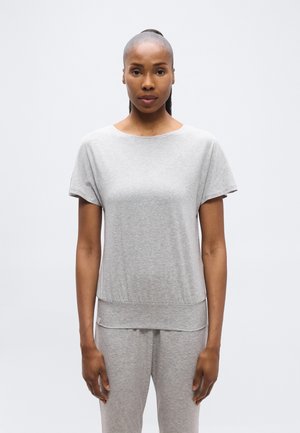 Femme aux longs cheveux attachés en queue de cheval, portant un haut à manches courtes gris clair et un pantalon assorti, debout devant un fond blanc uni.