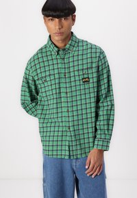 Green plaid shirt featuring check patterns, a button-down collar, and a small logo patch. Long sleeves, relaxed fit, and soft fabric.
