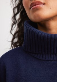 Tamsiai mėlynas megztinis su storu, raukšlėtu apykakle ir minkšta, tekstūrinėmis savybėmis, sutelkiant dėmesį į apykaklės sritį ir subtilius drabužio detalių bruožus.