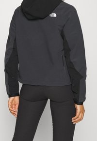 Sort vandtæt jakke med hætte, med logo på venstre skulder, lynlåslomme i siden og kontrasterende sorte detaljer på ærmerne.