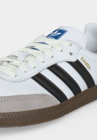 Weißer Adidas Samba Sneaker mit schwarzen Streifen, brauner strukturierter Sohle, weißen Schnürsenkeln, grauer Wildlederspitze und blauem Logo auf der Zunge.