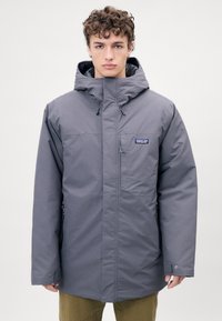 Veste grise à capuche avec une fermeture éclair sur le devant et des poches latérales. Fabriquée en matériau lisse et résistant à l'eau ; elle dispose d'un patch logo sur la poitrine.