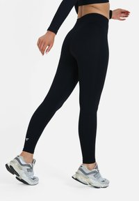 Black ribbed leggings with a high waist, featuring a seamless design and a small white logo at the ankle. Paired with gray athletic shoes.