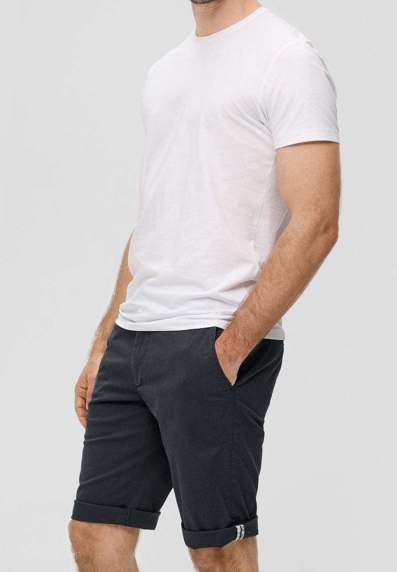 Homme portant un t-shirt blanc uni à manches courtes et un short noir retroussé, les mains dans les poches, sur un fond neutre.